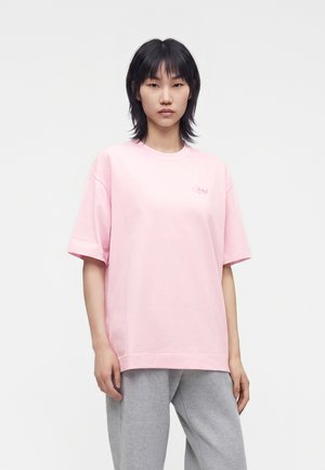 Jeune femme aux cheveux noirs portant un t-shirt ample rose clair et un pantalon gris, debout sur un fond blanc.