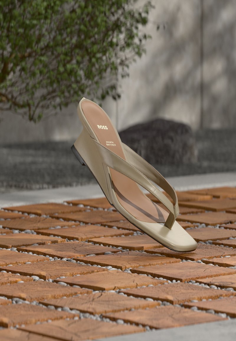 Πέδιλο με δερμάτινο λουράκι σε μπεζ χρώμα και λογότυπο BOSS στην εσωτερική σόλα, αιωρούμενο πάνω από ξύλινο πάτωμα με ξύλινες σανίδες και μικρές πέτρες ανάμεσά τους.
