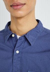 Camisa azul de botones con una tela texturada, un cuello puntiagudo y botones blancos. Cuenta con un bolsillo en el pecho. Vista en primer plano desde el cuello hacia abajo.