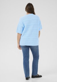 Suéter de punto azul claro con patrones texturizados, mangas cortas y un corte ancho, combinado con vaqueros azul medio y zapatos negros.