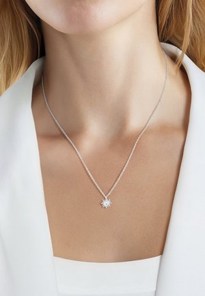 Collana con ciondolo argentato a forma di sole indossata da donna dalla pelle chiara e capelli biondi, con top bianco e blazer.