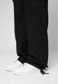 Person, der schwarze Hosen und weiße Schnürsenkelschuhe trägt, auf einem schlichten hellen Hintergrund steht.
