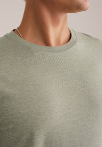 Prim-plan al unei persoane care poartă un tricou cu guler rotund de culoare verde deschis și un lănțișor subțire de argint, arătând gâtul și partea superioară a pieptului.