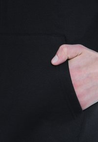 Poche d'un sweat à capuche noir avec une main partiellement insérée. Le matériau est doux, avec une texture côtelée et un design propre et simple.
