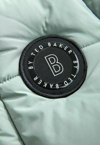 Close-up van een zwarte ronde Ted Baker logo patch met een witte "B" op lichtgroene gewatteerde stof.