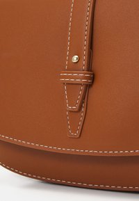 Bruine leren tas met witte stiksels, voorzien van een riem die vastzit met een goudkleurige stud. Gladde textuur en minimalistisch ontwerp.