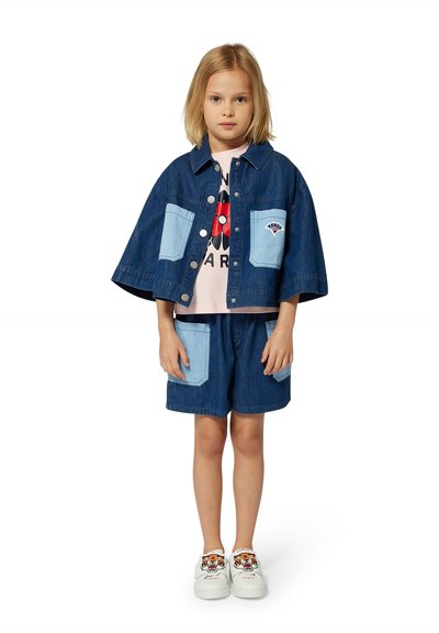 Denim jack met lichtblauwe patchzakken, gecombineerd met denim shorts met bijpassende zakken. Witte sneakers met een tijgerontwerp.