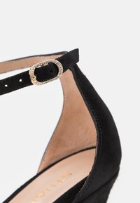 Talons en daim noir avec bride de cheville réglable ornée d'une boucle dorée embellie de strass transparents. Semelle intérieure beige avec marquage doré.