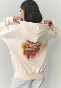 Šviesiai rožinė gobtuvėlė su dideliu gėlių grafiku ant nugaros ir baltu tekstu "FLEURISTE FLOWER SHOW", tekstūruota medžiaga, laisvas fasonas.