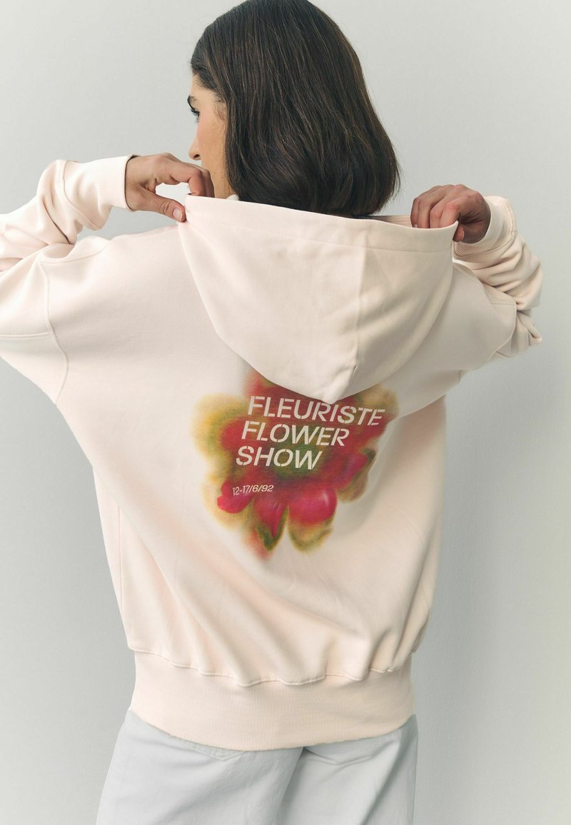 Šviesiai rožinė gobtuvėlė su dideliu gėlių grafiku ant nugaros ir baltu tekstu "FLEURISTE FLOWER SHOW", tekstūruota medžiaga, laisvas fasonas.