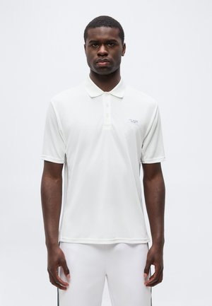 Muž v bielom polo tričku s krátkym rukávom a bielych nohaviciach, stojaci na jednoduchom svetlom pozadí, smerujúci dopredu.