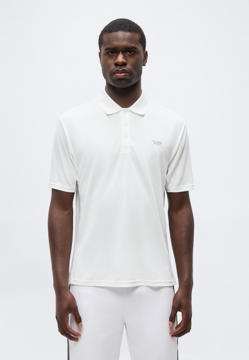 Άνδρας που φοράει λευκό πόλο με κοντά μανίκια και λευκό παντελόνι, στέκεται σε απλό φωτεινό φόντο, κοιτάζοντας μπροστά.