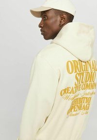 Sudadera color crema con un gran texto gráfico amarillo en la espalda. Presenta una capucha con cordones y una tela suave y lisa. Combinada con una gorra a juego.