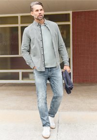 Man wandelt buiten gekleed in een licht overhemd met patroon, een grijs T-shirt, versleten blauwe spijkerbroek, witte sneakers, en houdt een donker vest in zijn rechterhand.