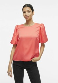 Blusa in satin corallo con maniche a puff e scollatura rotonda, abbinata a jeans neri. Tessuto morbido, fabbricazione leggera, design semplice.