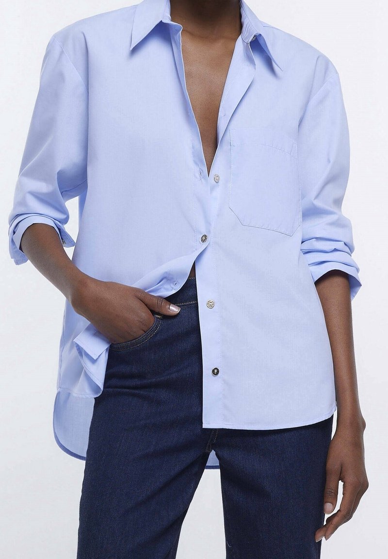 Ljusblå skjorta med knappar, bröstficka, uppvikta ärmar och rak nederkant. Tyget verkar slätt och något strukturerat.