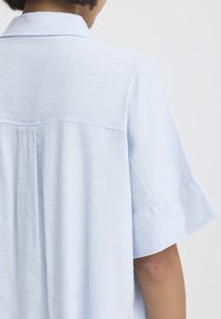 Personne portant une chemise à manches courtes bleu clair vue de dos, avec une coupe ample et un col, sur un fond blanc uni.