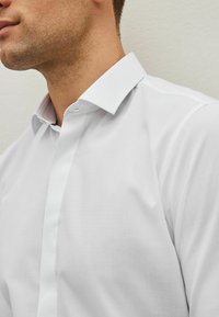 Mężczyzna w eleganckiej białej koszuli z klasycznym kołnierzykiem, widoczny od szyi do połowy klatki piersiowej na neutralnym tle.