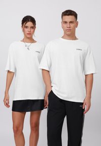 Camisetas blancas de gran tamaño con mangas cortas y cuello redondo, que presentan un pequeño logo negro en el pecho. Dos modelos se encuentran de pie uno al lado del otro.