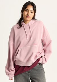 Light pink hooded sweatshirt with a front pocket, ribbed hem, and drawstrings. Notable branding in pink on the chest. Soft texture.