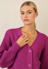 Paarse chunky gebreide cardigan met een V-hals, met gestructureerde strepen en twee grote knopen aan de voorkant. Zacht, comfortabel materiaal.