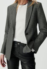 Czarna i biała marynarka w wzór houndstooth z klapami i zapięciem na jeden guzik, noszona na białej koszuli i czarnych spodniach.