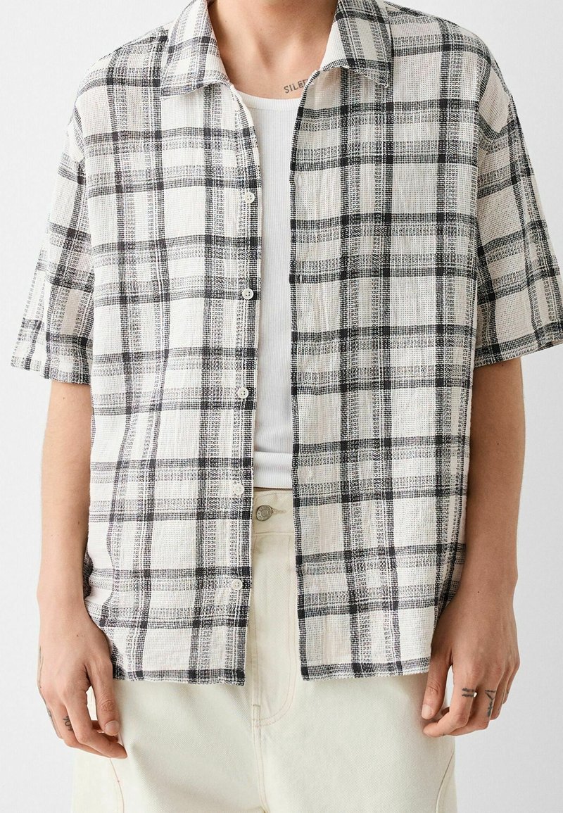 Personne portant une chemise à manches courtes à carreaux noir et blanc boutonnée sur un débardeur blanc côtelé et un pantalon de couleur claire.