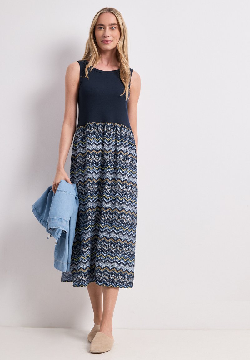 Femme en robe midi sans manches bleu marine à motifs tenant une veste bleu clair, debout devant un mur blanc uni, portant des chaussons beige.