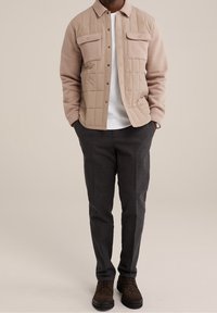 Beige vadderad skjortjacka med tryckknappar och två bröstfickor, matchad med mörkgrå strukturerade byxor och bruna snörskor.