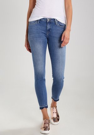 Žena v krátkých modrých úzkých džínách, bílém tílku a metalických růžově zlatých slip-on teniskách, stojící před jednoduchým pozadím.