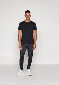 T-shirt de sport noir à manches courtes avec un logo, associé à un pantalon noir fuselé et des baskets blanches avec des accents verts.