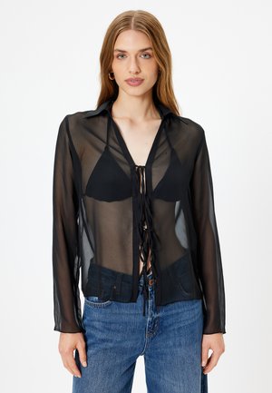 Blouse noire transparente avec manches longues, col et fermeture à cordon à l'avant. Associée à un top de bikini noir et un jean bleu.