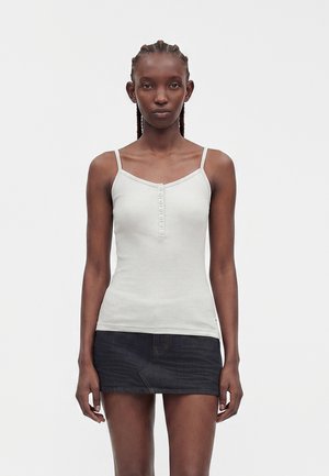 Kvinde med fletninger iført en hvid knappet camisole og et sort denim miniskørt, stående mod en ensfarvet hvid baggrund.