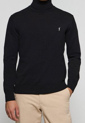 Man wearing black turtleneck sweater with small white polo player logo and beige pants, hands at sides.