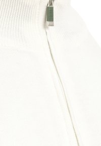 Pull en tricot blanc avec un col montant, doté d'une fermeture éclair argentée et d'une tirette vert clair. Texture lisse et bords côtelés.
