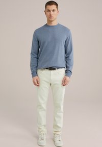 Gaiši zils adīts džemperis, krēmkrāsas bikses un baltas kedas. Modelis stāv pret neitrālu fona, rokas pie sāniem.