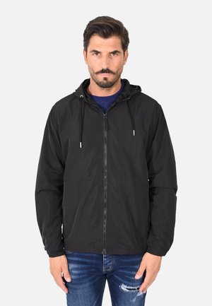 Uomo adulto con capelli scuri e barba che indossa una giacca nera con cappuccio e zip sopra un maglione blu navy e jeans strappati, in piedi davanti a uno sfondo semplice.