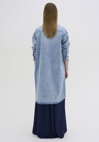 Abito camicia in denim con maniche lunghe, colore azzurro chiaro e orlo arrotondato, indossato sopra una gonna maxi blu navy. Tessuto morbido e vestibilità ampia.