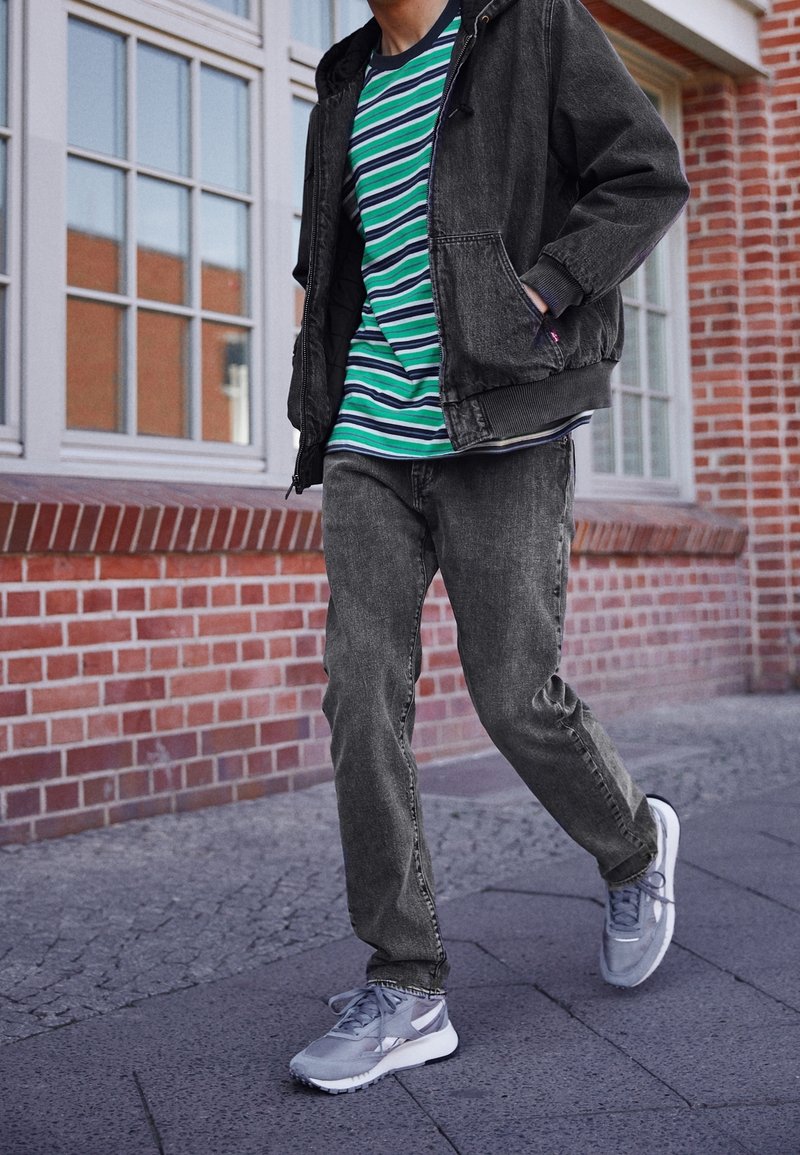 Avslappnad outfit med en svart denimjacka, grön och blå randig t-shirt, grå jeans och grå sneakers med vita accenter.