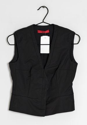Czarna bezrękawnik z głębokim dekoltem w kształcie litery V, dwoma przednimi kieszeniami, gładką fakturą i czerwoną metką przy szyi. Metka przymocowana sznurkiem.
