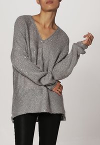 Harmaa puuvillane kudum V-kaelusega, pikkade varrukatega ja vabalt langev. Omab tekstureeritud mustrit ja peeneid litre, et lisada detaili.