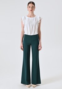 Donna in piedi con una blusa bianca senza maniche a volant, pantaloni palazzo verde scuro, scarpe beige a punta e orecchini a cerchio in argento.