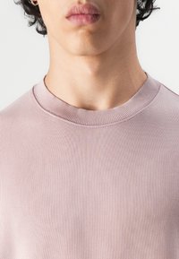 Lys rosa strikket t-skjorte med rund hals. Ribbestruktur langs kragen. Enklere, sømløs design uten synlige mønstre eller detaljer.