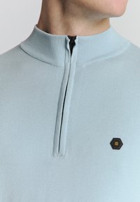 Lichtblauw gebreid pullover met een hoge kraag, voorzien van een halve rits en een hexagonaal logo-opnieuw in zwart en goud op de borst.