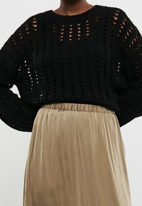 Schwarzer gehäkelter Pullover mit langen Ärmeln, kombiniert mit einem hochgeschnittenen, beigen Faltenrock mit elastischem Bund.