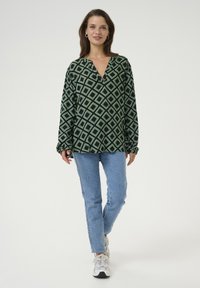 Blouse à motifs verts et noirs avec un col en V et des manches longues, associée à un jean bleu clair et des baskets grises.