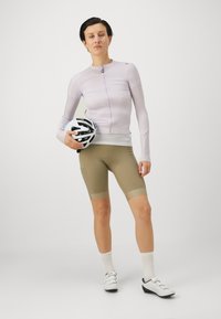Light purple, fitted cycling jersey with a front zip. Olive-green shorts with a chamois. Wearing a black and white cycling helmet.