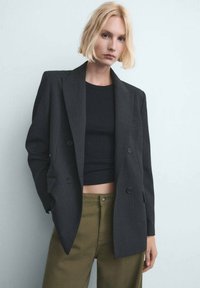 Persona rubia con una blazer negra de doble botonadura sobre un crop top negro y pantalones verde oliva, de pie contra una pared clara.