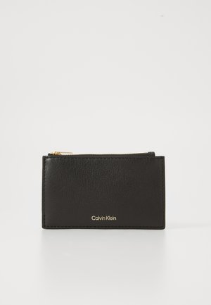 Porte-cartes en cuir noir Calvin Klein avec fermeture éclair dorée en haut et nom de la marque embossé en or à l'avant.