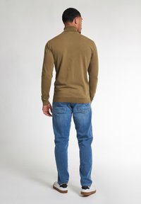 Olivově zelená polo tričko s dlouhým rukávem a žebrovaným límečkem, doplněno světle modrými džínami a bílými teniskami s černými akcenty.